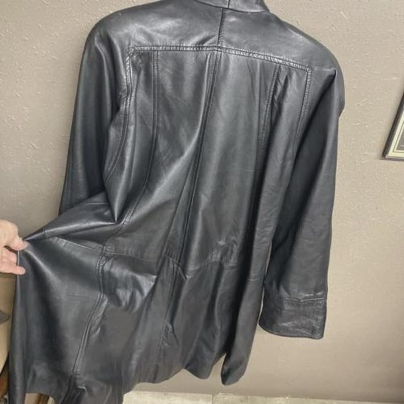 K&ouml;nnte beinhalten: Schwarze Lederjacke mit langer L&auml;nge und klassischem Design. Die Jacke hat einen Kragen, lange &Auml;rmel und einen Knopfverschluss. Das Leder scheint weich und geschmeidig zu sein, mit sichtbaren Nahtdetails.