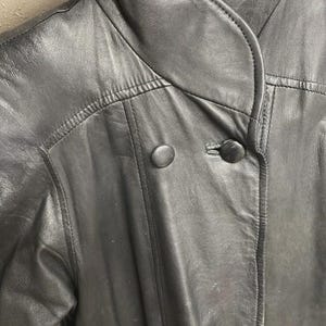 K&ouml;nnte beinhalten: Nahaufnahme einer schwarzen Lederjacke mit rundem Kragen und zwei gro&szlig;en, runden Kn&ouml;pfen. Die Jacke hat sichtbare Nahtdetails und ein klassisches Design, das auf einen Vintage- oder Retro-Stil hindeutet.