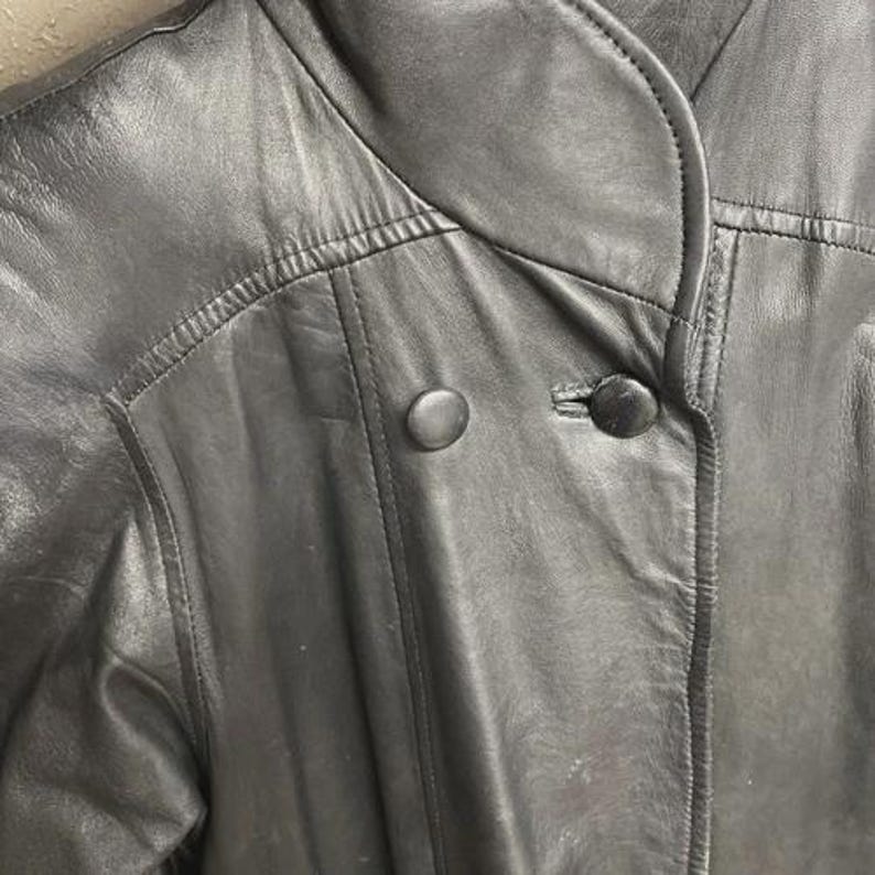 K&ouml;nnte beinhalten: Nahaufnahme einer schwarzen Lederjacke mit rundem Kragen und zwei gro&szlig;en, runden Kn&ouml;pfen. Die Jacke hat sichtbare Nahtdetails und ein klassisches Design, das auf einen Vintage- oder Retro-Stil hindeutet.