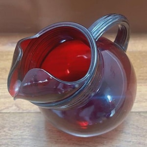 Könnte beinhalten: Ein rubinroter Glaskrug mit rundem Körper und gebogenem Ausguss. Der Griff ist gerippt und der Krug hat eine glänzende Oberfläche. Der Krug wird wahrscheinlich zum Servieren von Getränken verwendet.