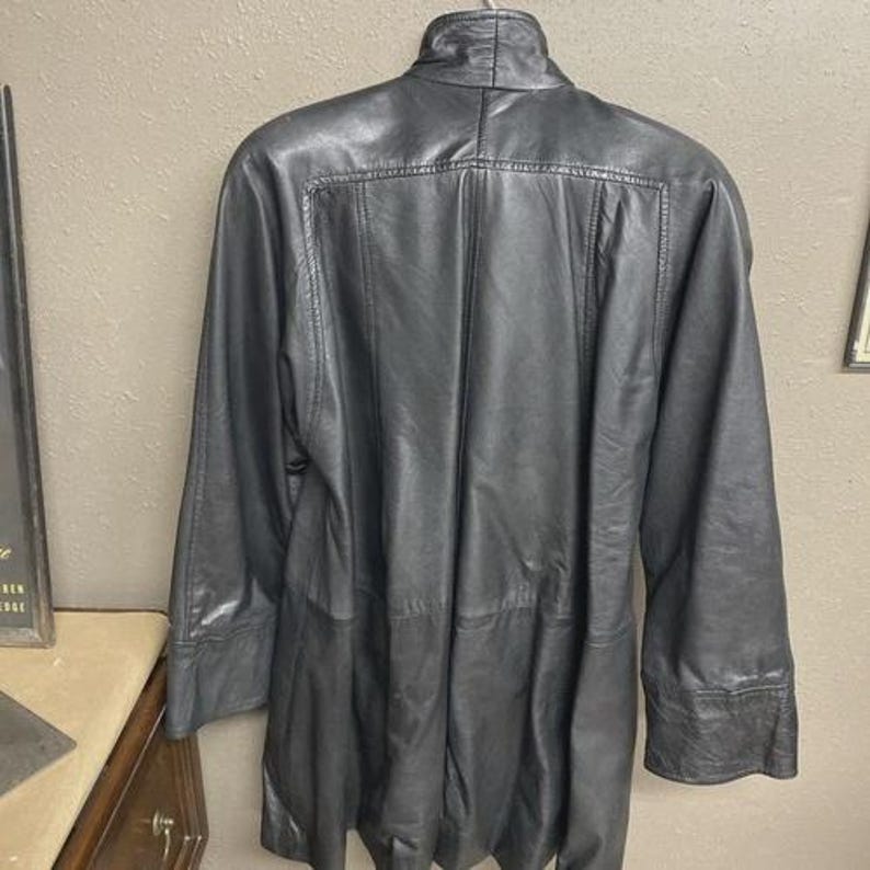 K&ouml;nnte beinhalten: Eine schwarze Lederjacke mit hohem Kragen und langen &Auml;rmeln. Die Jacke hat eine taillierte Passform und ein knielanges Design. Die R&uuml;ckseite der Jacke hat vertikale N&auml;hte und eine subtile Textur. Die Jacke h&auml;ngt an einer Wand.