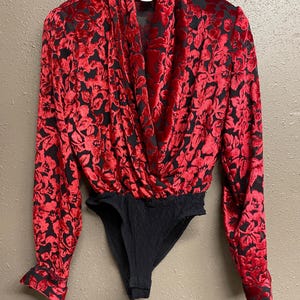 May include: A red floral-patterned bodysuit with long sleeves and a draped neckline. The fabric has a velvet texture, and the bottom portion is black. The garment is hanging on a black hanger.