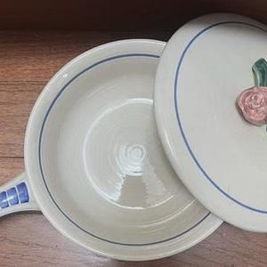 May include: Off-white ceramic casserole dish with a lid. The dish has a handle with blue and white squares. The lid features a pink rose and green leaves. A blue line borders the rim of the dish and the lid.