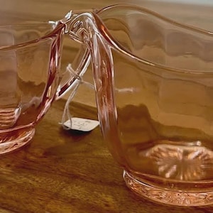 Op de afbeelding: Twee roze glazen serveerschalen. De ene is een kleine kom met een handvat, en de andere is een grotere roomkan met een handvat en een tuit. Beide hebben een decoratief patroon op de bodem en zijn gemaakt van transparant glas.