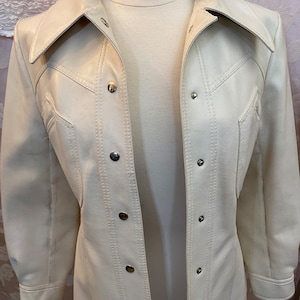 Könnte beinhalten: Eine weiße Kunstlederjacke mit Knopfleiste und langen Ärmeln. Die Jacke hat einen spitzen Kragen und eine leicht taillierte Silhouette.