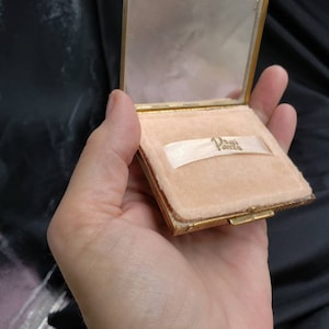 以下が含まれることがあります： 鏡とピンクのベルベットの内装が付いた金のコンパクト。コンパクトには、「D'Orsay Paris」という文字が印刷された白いリボンが付いています。