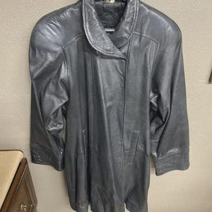 K&ouml;nnte beinhalten: Ein dunkelgrauer Ledermantel mit rundem Kragen und langen &Auml;rmeln. Der Mantel hat einen Knopfverschluss und zwei Vordertaschen. Das Leder scheint weich zu sein und hat einen leichten Glanz. Der Mantel h&auml;ngt an einem Kleiderb&uuml;gel.