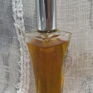 May include: A clear glass perfume bottle with a silver cap. The bottle is filled with a golden-colored liquid. The bottle has a unique, angular shape. The background is a sheer, white fabric.