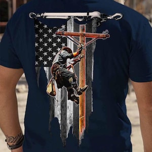 Könnte beinhalten: Marineblaues T-Shirt mit einer Grafik eines Elektrikers, der einen Strommast erklimmt, vor einer verwitterten amerikanischen Flagge. Das Design enthält eine detaillierte Illustration des Elektrikers und des Masts.