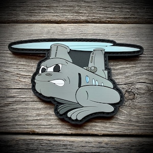 May include: A gray cartoon helicopter with a grumpy expression, with a black outline and a blue rotor, is depicted on a black patch.