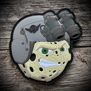 May include: A gray and green patch with a cartoon face wearing a helmet with night vision goggles. The face has a large, toothy grin and a textured, cratered surface.