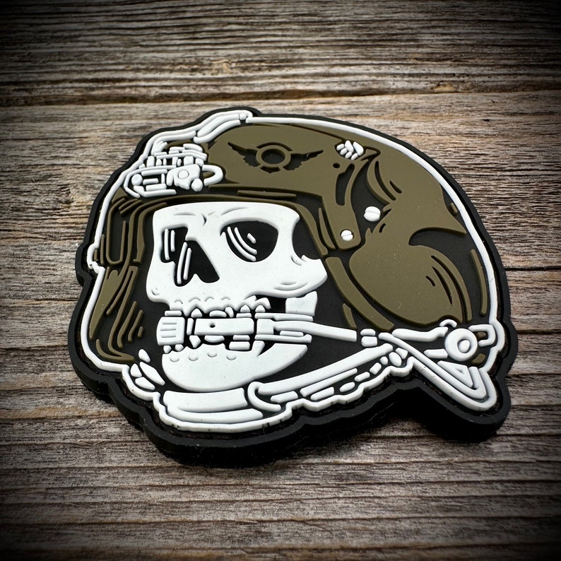 May include: A white skull wearing a green military helmet with a black outline. The skull is wearing a breathing apparatus and has a detailed design.