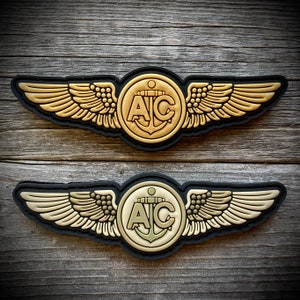 May include: Two gold-colored embroidered patches with black outlines. Each patch features a pair of wings with a circle in the center. The circle contains the letters "A/C" and an anchor. The top patch has a lighter gold color and the bottom patch has a darker gold color.