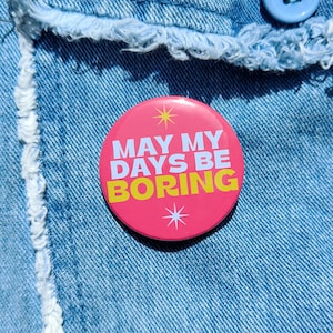 May include: Pink button with white text that reads "May My Days Be Boring" in yellow letters. The button has a white star design on either side of the text.