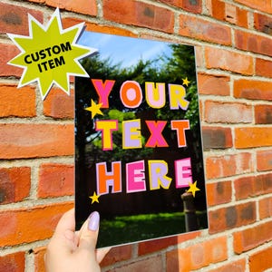 以下が含まれることがあります： カラフルで太字の文字で「YOUR TEXT HERE」と書かれた、黒色の反射性カスタムサイン。左上には「CUSTOM ITEM」と書かれた黄色のスターバーストステッカーがあります。レンガの壁を背景に、サインが掲げられています。