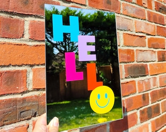 May include: A rectangular mirror with the word "HELLO" in bright, colorful letters. The letters are blue, pink, purple, and orange. A yellow smiley face is below the word "HELLO". The mirror is held in front of a brick wall.