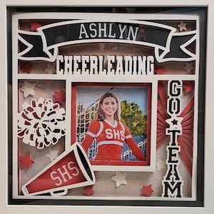 May include: A white frame with a red, white, and black design. The frame includes a photo of a girl in a red and white cheerleading uniform, a megaphone, a pom-pom, and the words "ASHLYN CHEERLEADING GO TEAM".