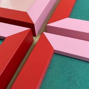 May include: Close-up of several picture frames in shades of red and pink. The frames are arranged at angles, with the text "CHAI ME UP" visible on one frame. The background is a teal colour.