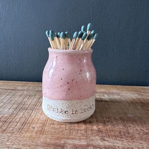 May include: A pink ceramic match holder with the text "Strike it lucky" on the base. The holder is filled with wooden matches with blue tips. The holder sits on a wooden surface.