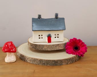 May include: A small white ceramic house with a gray roof and a red door. The house is sitting on a wooden base. There is a red mushroom and a pink flower in the background.