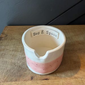 May include: A white ceramic bag and spoon holder with a pink band around the middle. The words "Bag & Spoon" are printed on the top rim. The holder is sitting on a wooden surface.