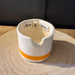 May include: White ceramic dish with a yellow stripe around the rim. The dish has the words "Bag & Spoon" written on the inside.