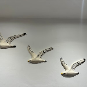 Op de afbeelding: Drie witte keramische meeuwen wanddecoraties met zwarte vlekken. De meeuwen zijn in de vlucht en kijken in verschillende richtingen.