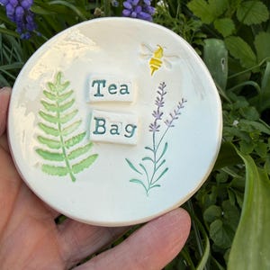 Könnte beinhalten: Ein runder, cremefarbener Keramik-Teebeutelhalter mit den grünen Worten "Tea Bag". Der Halter zeigt einen Farn, einen Lavendelzweig und eine gelbe Biene. Umgeben von grünem Laub.