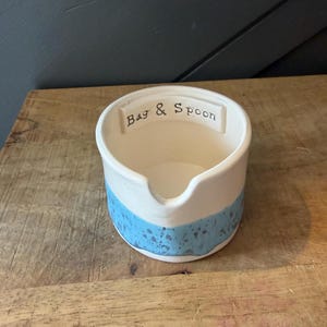 Op de afbeelding: Een witte keramische zak- en lepelhouder met een blauwe textuurband rond de basis. De woorden "Bag & Spoon" staan in het zwart op de bovenrand. De houder heeft een tuit.
