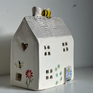 May include: A white ceramic house-shaped candle holder with a heart-shaped window and square windows. It features a bee and flower design, a chimney, and a yellow and black bee figurine on the roof.