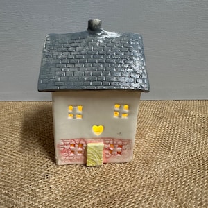 Peut inclure: Une petite maison en céramique avec un toit en briques grises, des murs blancs et une base en briques roses. La maison a deux fenêtres avec de la lumière qui brille à travers elles et une découpe en forme de cœur jaune au-dessus de la porte.