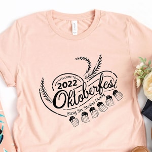 May include: A pink t-shirt with a black graphic that says "Welcome to 2022 Oktoberfest Stay Six Beers Ready". The graphic features a circle with a wheat stalk design and beer mugs.