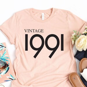 May include: A light pink t-shirt with the text "VINTAGE 1991" in black. The shirt is styled with dark wash jeans, white sandals, a floral clutch, sunglasses, and a gold bracelet. A bouquet of flowers is also included.