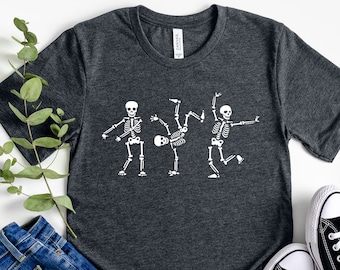 Könnte beinhalten: Ein dunkelgraues T-Shirt mit einer weißen Grafik von drei tanzenden Skeletten. Die Skelette sind in verschiedenen Posen, wobei ein Skelett einen Rückwärtssalto macht.