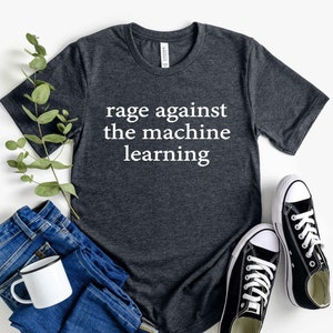 May include: A dark gray t-shirt with the text "rage against the machine learning" in white lettering. The shirt is laid flat on a white background with a pair of black canvas shoes and a pair of blue jeans.