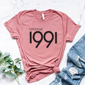May include: A dusty rose-colored t-shirt with the text "VINTAGE 1991" in black. The shirt is paired with light blue denim jeans with distressed details. A sprig of green leaves is placed to the left of the shirt.