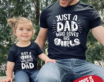May include: A black t-shirt with white text that reads "Just a Dad who loves his Girls". A smaller black t-shirt with white text that reads "Just a Girl who loves her Dad" is worn by a young girl. The girl is sitting on a wall with a man wearing the larger t-shirt.