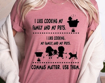 Può includere: Una maglietta rosa con una grafica nera che dice "Mi piace cucinare la mia famiglia e i miei animali domestici." La grafica include anche una pentola con del vapore che esce, una famiglia di tre persone e un cane. Il testo "Mi piace cucinare, la mia famiglia e i miei animali domestici." è anche incluso nella grafica. La grafica termina con il testo "Le virgole contano. Usale."