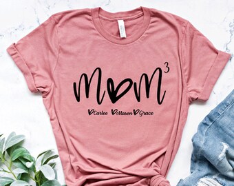 May include: A pink t-shirt with the word "MOM" in black script with a heart in the "O". Below the word "MOM" is the number "3" and the names "Carlee", "Mason", and "Grace" with hearts in front of each name.