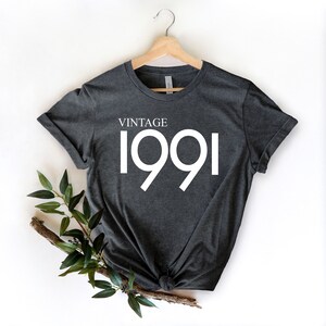 May include: Dark gray t-shirt with the text "VINTAGE 1991" in white. The shirt is hanging on a wooden hanger against a white background. A sprig of greenery is placed at the bottom left.