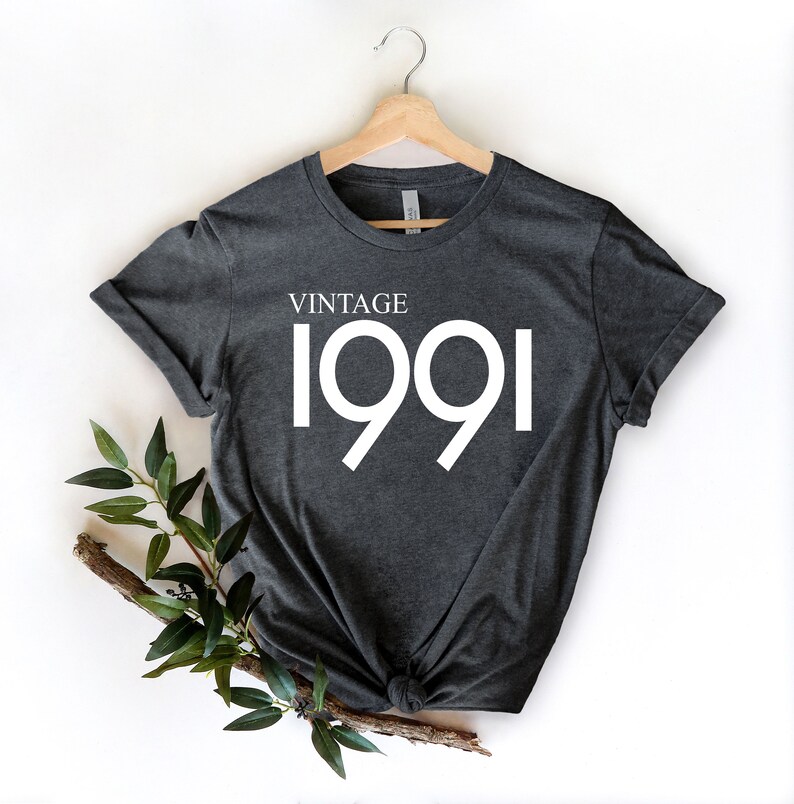 May include: Dark gray t-shirt with the text "VINTAGE 1991" in white. The shirt is hanging on a wooden hanger against a white background. A sprig of greenery is placed at the bottom left.