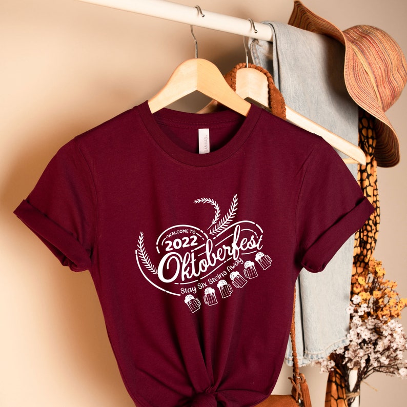 May include: Burgundy t-shirt with a white graphic that says "Welcome to 2022 Oktoberfest Stay Six Feet Away". The graphic also includes a beer stein and wheat.