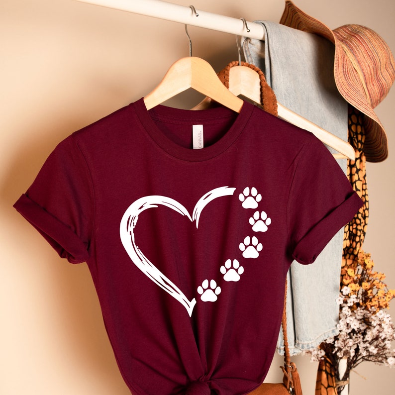 May include: A maroon t-shirt with a white heart outline and paw prints. The heart is on the left side of the shirt and the paw prints are on the right side of the shirt.