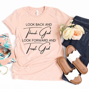 May include: A peach-colored t-shirt with the text "Look back and thank God, look forward and trust God" in black cursive lettering.