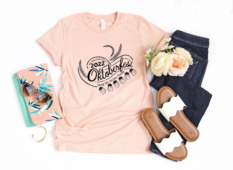 May include: A pink t-shirt with a black graphic that says "Welcome to 2022 Oktoberfest Stay Six Beers Ready". The graphic features a circle with a wheat stalk design and beer mugs.