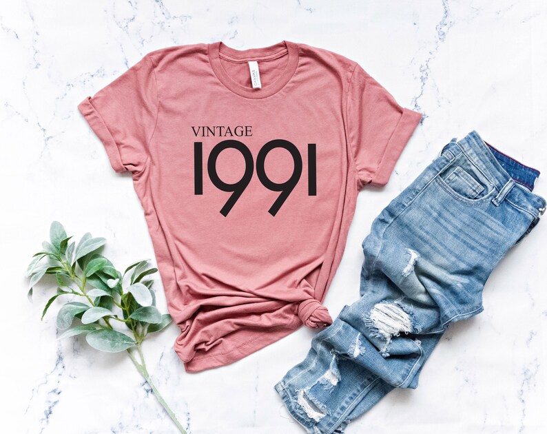 May include: A dusty rose-colored t-shirt with the text "VINTAGE 1991" in black. The shirt is paired with light blue denim jeans with distressed details. A sprig of green leaves is placed to the left of the shirt.