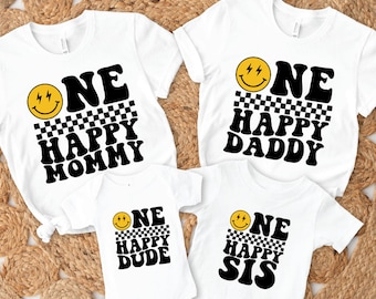 May include: Four white t-shirts with black text and a yellow smiley face graphic. The shirts say "One Happy Mommy", "One Happy Daddy", "One Happy Dude", and "One Happy Sis".