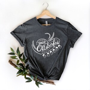 May include: A dark gray t-shirt with a white graphic design that says "Welcome to 2022 Oktoberfest Stay Six Steins Away" with a graphic of wheat stalks and beer steins.