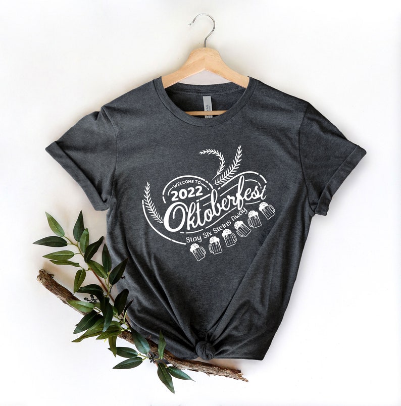 May include: A dark gray t-shirt with a white graphic design that says "Welcome to 2022 Oktoberfest Stay Six Steins Away" with a graphic of wheat stalks and beer steins.