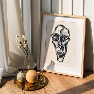 Può includere: Un ritratto in bianco e nero incorniciato di un volto con un naso lungo e un'espressione cupa. L'opera d'arte è esposta su un pavimento in legno, accanto a un vassoio dorato con sfere decorative e un vaso di fiori bianchi. La cornice è in legno chiaro.